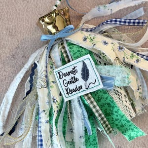 May include: A decorative tassel featuring a gold-colored crown, blue ribbon, and a variety of fabric strips in white, green, and blue floral and gingham patterns. A small square tag reads "Dearest Gentle Reader".