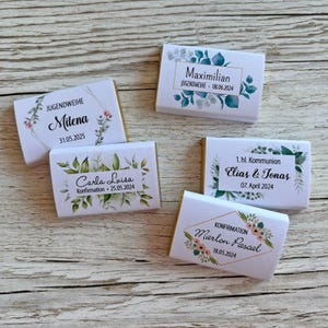 Op de afbeelding: Vijf witte rechthoekige chocoladerepen met gepersonaliseerde tekst en bloemmotieven. De repen zijn gelabeld met namen en data voor evenementen zoals Jugendweihe, Vormsel en Eerste Communie. Gerangschikt op een houten oppervlak.