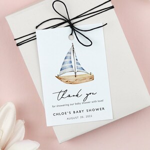 May include: A white gift tag with a black string tied around it. The tag features a watercolor illustration of a sailboat and the text "thank you for showering our baby shower with love! Chloe's Baby Shower August 20, 2021".
