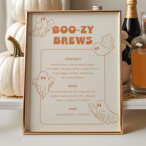 Könnte beinhalten: Ein goldgerahmtes Schild mit den Worten "BOO-ZY BREWS" und einer Liste von Cocktails, Bieren und Weinen. Das Schild ist mit Geisterillustrationen verziert. Weiße Kürbisse und Alkoholflaschen sind um das Schild auf einem weißen Tablett angeordnet.