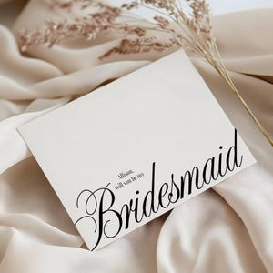 May include: A white card with the words "Allison, will you be my Bridesmaid" in black script. The card is set against a backdrop of cream-coloured fabric and dried floral accents.