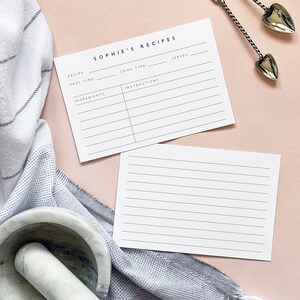 May include: Two blank recipe cards with lines for writing down recipes. The cards are on a pink surface with a gray and white striped towel and a white mortar and pestle.