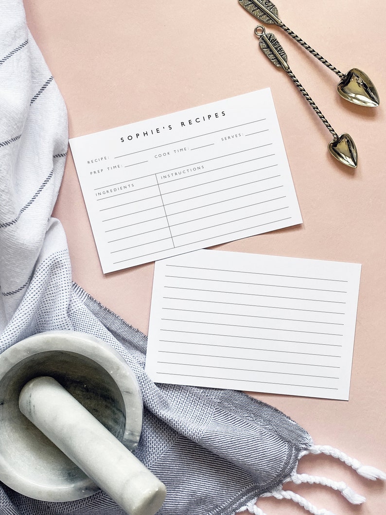 May include: Two blank recipe cards with lines for writing down recipes. The cards are on a pink surface with a gray and white striped towel and a white mortar and pestle.