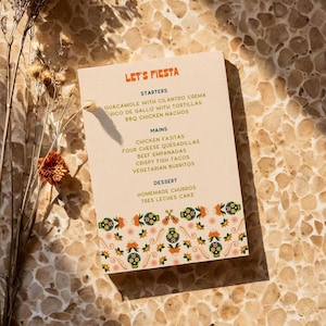 Może przedstawiać: Menu zatytułowane "Let's Fiesta" oferujące wybór meksykańskich potraw, w tym przystawki, dania główne i desery. Menu jest ozdobione kolorową ramką z cukrowymi czaszkami i wzorami kwiatowymi.