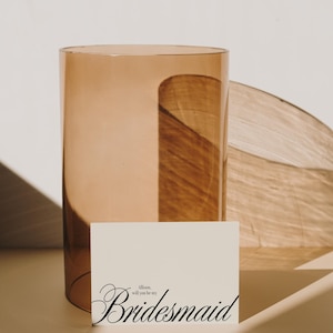 May include: A tall, amber-toned glass vase with a curved design, next to a card that reads "Allison, will you be my Bridesmaid" in elegant script. The vase is cylindrical, and the card is a soft cream colour.