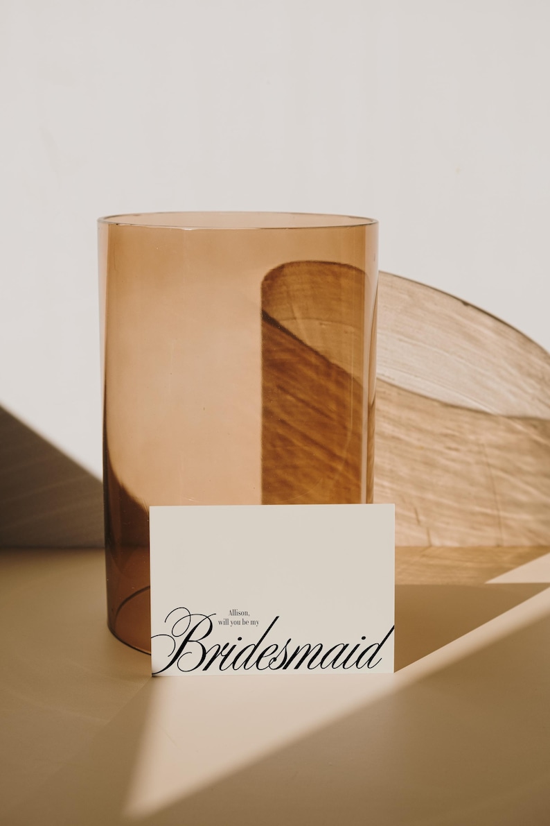 May include: A tall, amber-toned glass vase with a curved design, next to a card that reads "Allison, will you be my Bridesmaid" in elegant script. The vase is cylindrical, and the card is a soft cream colour.