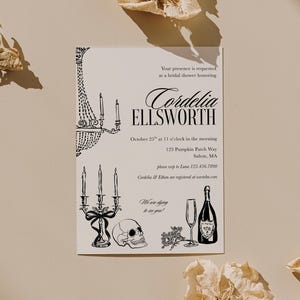 May include: A white rectangular bridal shower invitation card with black ink illustrations of a chandelier, a candelabra, a skull, a champagne glass, and a bottle. The text reads: Your presence is requested at a bridal shower honoring Cordelia ELLSWORTH, October 25th at 11 o'clock in the morning, 123 Pumpkin Patch Way, Salem, MA, please rsvp to Luna 123.456.7890, Cordelia & Ethan are registered at westelm.com, We are dying to see you!