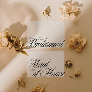 May include: Two white cards with elegant black script reading "Bridesmaid" and "Maid of Honor". Dried flowers in beige and brown are scattered around the cards on a neutral background. The text "Allison, will you be my" is also present.
