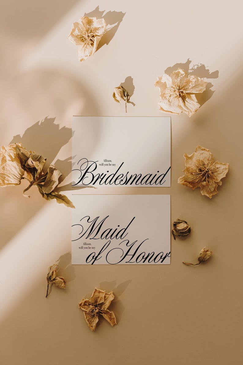 May include: Two white cards with elegant black script reading "Bridesmaid" and "Maid of Honor". Dried flowers in beige and brown are scattered around the cards on a neutral background. The text "Allison, will you be my" is also present.