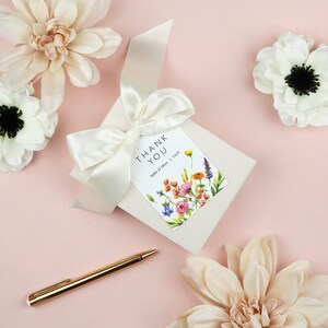 May include: A white card with a floral design and the text "Thank You" with the names "Sophie & Oliver" and the date "1.5.23". The card is tied with a white satin ribbon and surrounded by white flowers on a pink background.