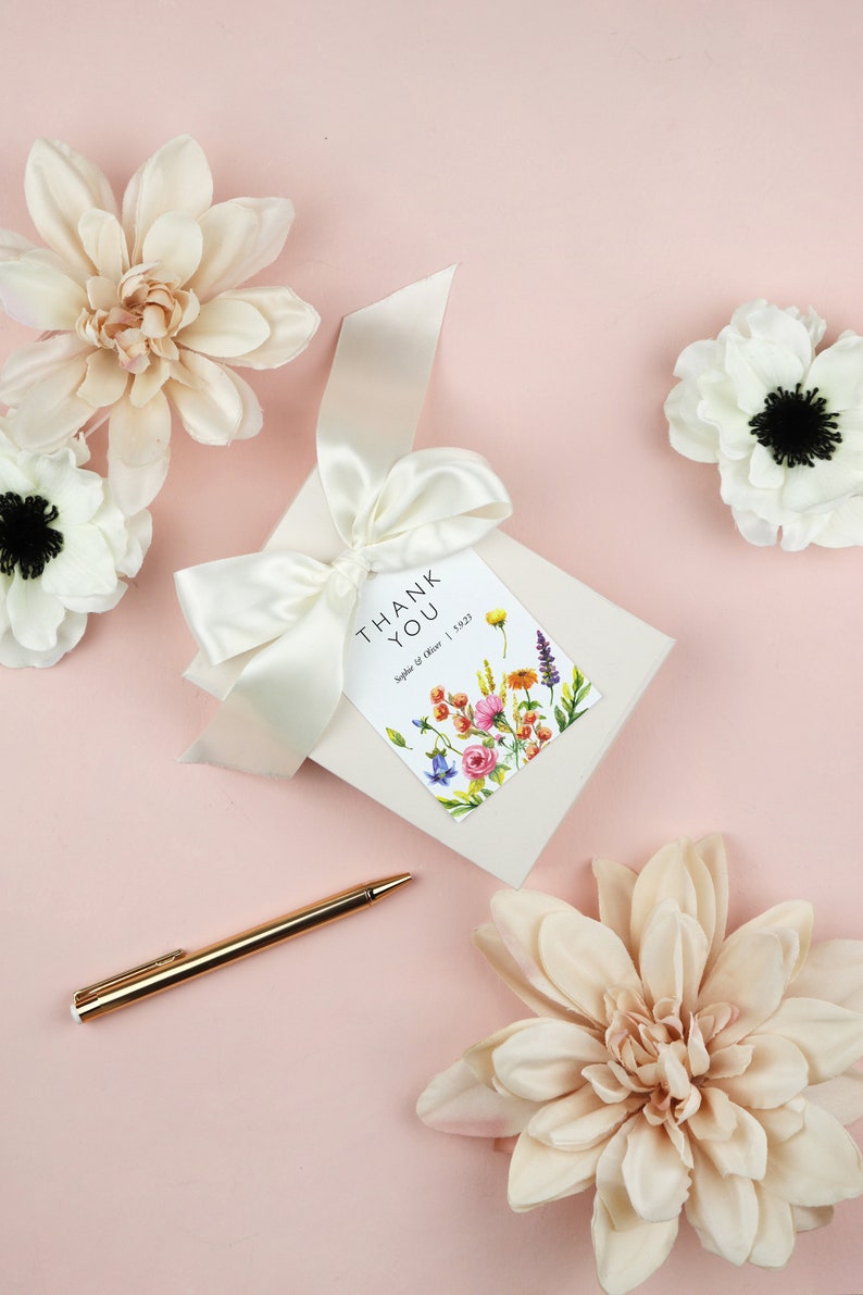 May include: A white card with a floral design and the text "Thank You" with the names "Sophie & Oliver" and the date "1.5.23". The card is tied with a white satin ribbon and surrounded by white flowers on a pink background.