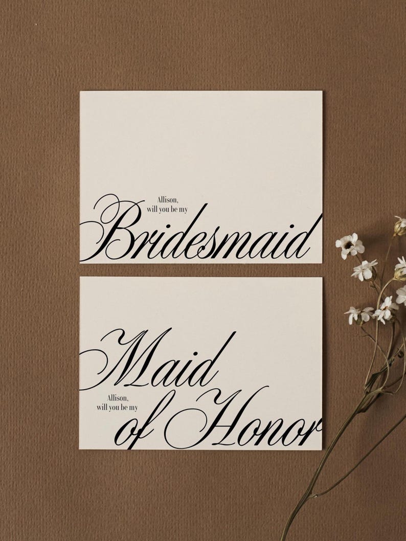May include: Two ivory cards with black calligraphy. The top card reads "Bridesmaid" with the text "Allison, will you be my" above it. The bottom card says "Maid of Honour" with the same preceding text. A sprig of white flowers is in the background.