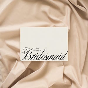 May include: A cream-coloured card with the word "Bridesmaid" in elegant black script. The text "Allison, will you be my" is above the main word. The card rests on a beige, draped fabric background.