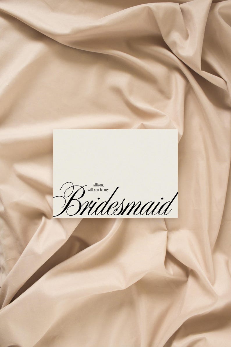 May include: A cream-coloured card with the word "Bridesmaid" in elegant black script. The text "Allison, will you be my" is above the main word. The card rests on a beige, draped fabric background.
