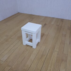 Puede incluir: Un taburete de madera blanco con una tapa cuadrada y un diseño de marco simple y abierto. El taburete está sentado en un suelo de madera marrón claro.