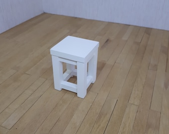 Könnte beinhalten: Ein weißer Holzhocker mit quadratischer Oberseite und einem einfachen, offenen Gestell. Der Hocker steht auf einem hellbraunen Holzboden.