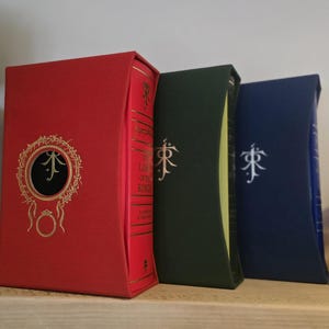 Pode incluir: Tr&ecirc;s livros de capa dura com capas vermelha, verde e azul. Os livros s&atilde;o todos do mesmo tamanho e t&ecirc;m um design semelhante. As capas apresentam um s&iacute;mbolo estilizado e o t&iacute;tulo "O Senhor dos An&eacute;is" no livro vermelho.