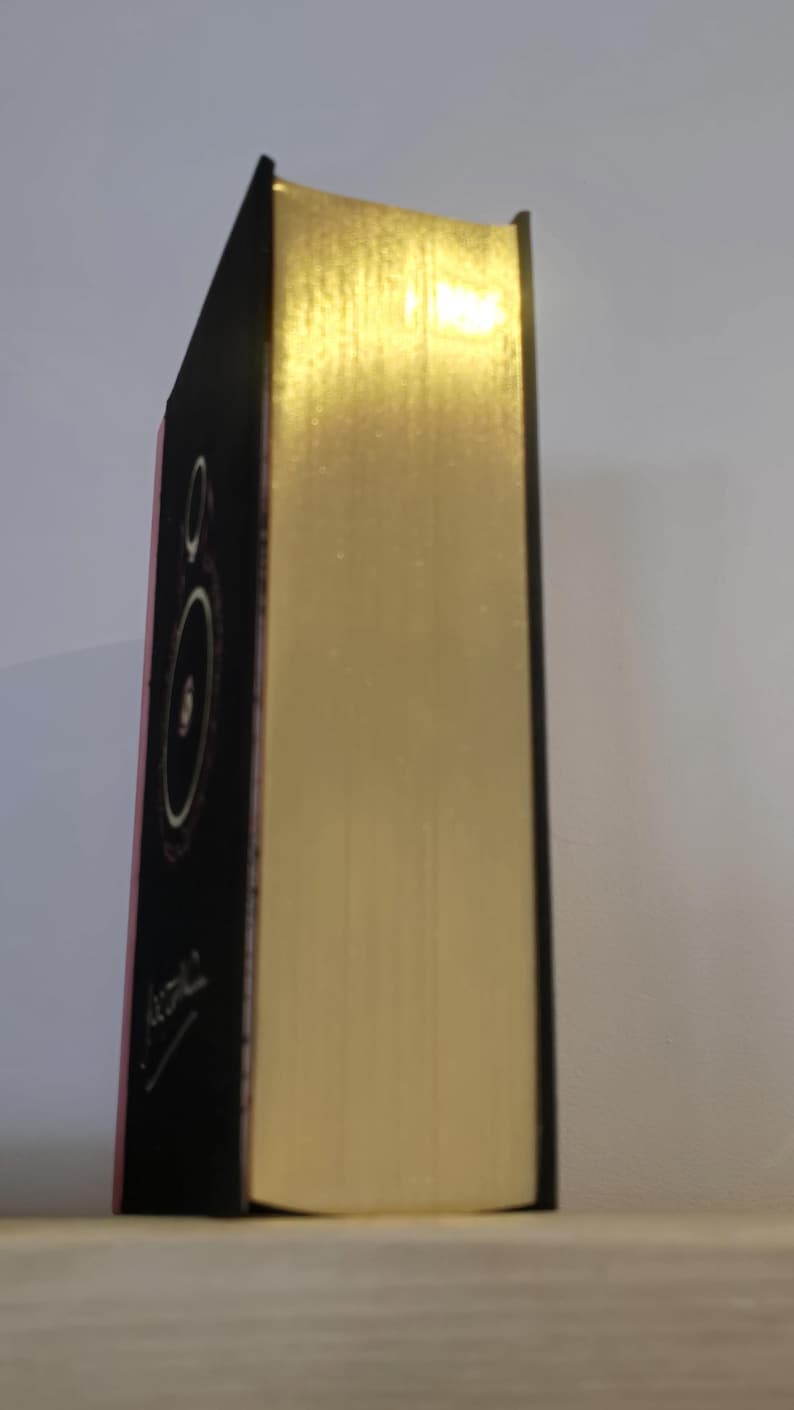 Pode incluir: Um livro encadernado em preto com uma lombada dourada e um design circular na capa frontal. O livro &eacute; intitulado "Atomic" em branco.