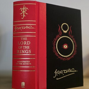 Pode incluir: Um livro encadernado em vermelho com letras douradas intitulado "O Senhor dos An&eacute;is" ilustrado pelo autor, J.R.R. Tolkien. O livro tem uma lombada preta com uma ilustra&ccedil;&atilde;o dourada do Olho de Sauron.