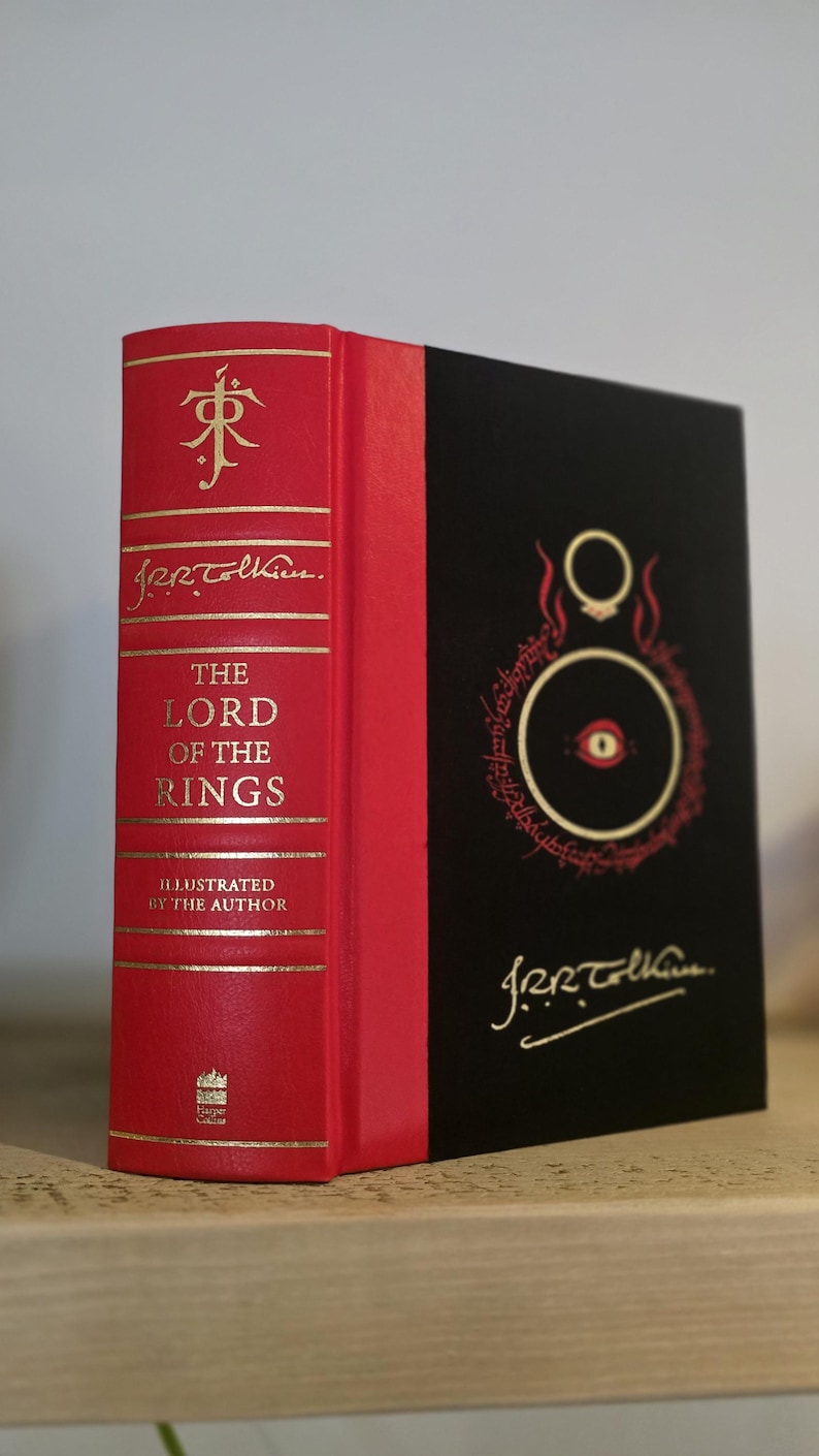 Pode incluir: Um livro encadernado em vermelho com letras douradas intitulado "O Senhor dos An&eacute;is" ilustrado pelo autor, J.R.R. Tolkien. O livro tem uma lombada preta com uma ilustra&ccedil;&atilde;o dourada do Olho de Sauron.