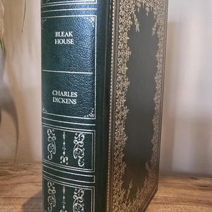 Pode incluir: Um livro de capa dura verde escuro intitulado "Bleak House" de Charles Dickens. A capa apresenta detalhes dourados e texto. A lombada mostra desenhos e o título e autor. O livro está sobre uma superfície de madeira.