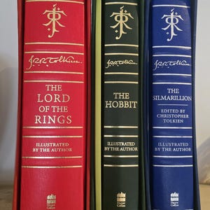 Pode incluir: Tr&ecirc;s livros encadernados com letras douradas e ilustra&ccedil;&otilde;es. Os livros s&atilde;o intitulados "O Senhor dos An&eacute;is", "O Hobbit" e "O Silmarillion".