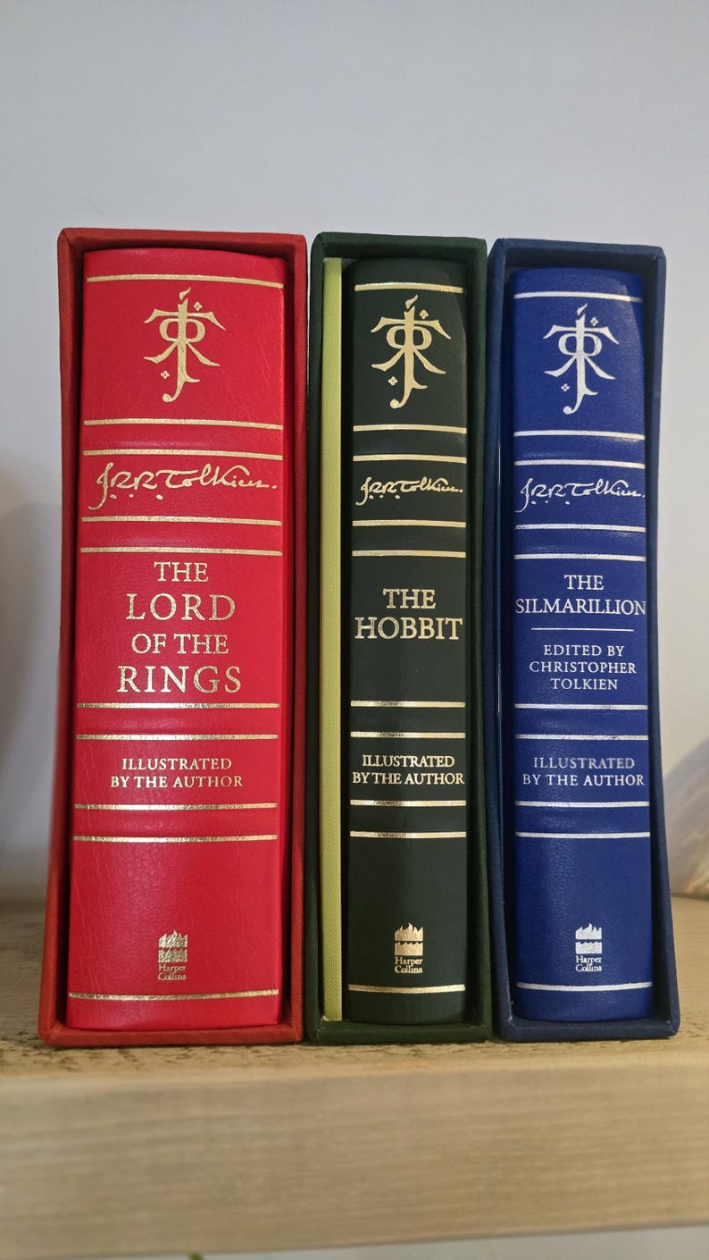 Pode incluir: Tr&ecirc;s livros encadernados com letras douradas e ilustra&ccedil;&otilde;es. Os livros s&atilde;o intitulados "O Senhor dos An&eacute;is", "O Hobbit" e "O Silmarillion".