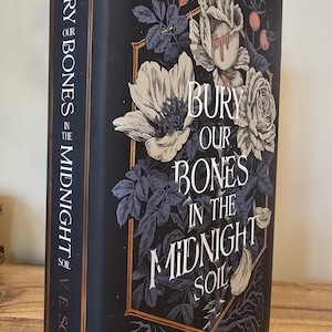 May include: A hardback book titled "Bury Our Bones in the Midnight Soil" by V.E. Schwab. The cover is black with floral illustrations in white and grey, and the spine features the title and author's name in white.