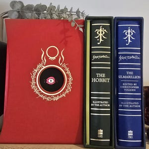 Pode incluir: Tr&ecirc;s livros encadernados com letras douradas nas lombadas. O livro &agrave; esquerda &eacute; vermelho com um c&iacute;rculo preto e um olho branco no centro. O livro do meio &eacute; verde com o t&iacute;tulo "O Hobbit" e o autor "J.R.R. Tolkien". O livro &agrave; direita &eacute; azul com o t&iacute;tulo "O Silmarillion" e o autor "J.R.R. Tolkien".