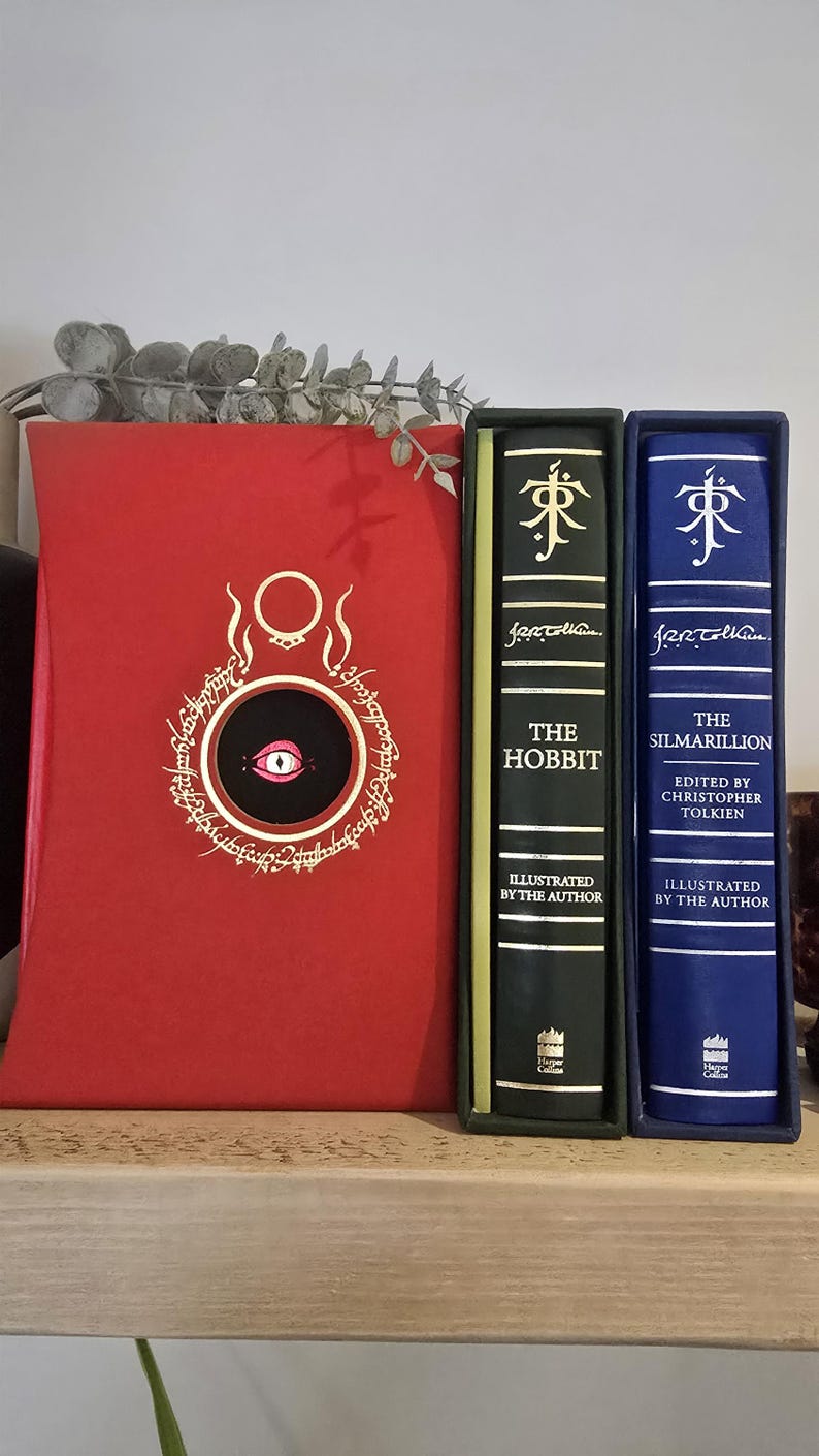 Pode incluir: Tr&ecirc;s livros encadernados com letras douradas nas lombadas. O livro &agrave; esquerda &eacute; vermelho com um c&iacute;rculo preto e um olho branco no centro. O livro do meio &eacute; verde com o t&iacute;tulo "O Hobbit" e o autor "J.R.R. Tolkien". O livro &agrave; direita &eacute; azul com o t&iacute;tulo "O Silmarillion" e o autor "J.R.R. Tolkien".
