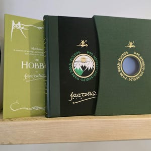 Pode incluir: Um livro de capa dura verde com o t&iacute;tulo "O Hobbit" de J.R.R. Tolkien. O livro est&aacute; em uma caixa deslizante com uma capa verde e um recorte circular que revela um fundo azul.