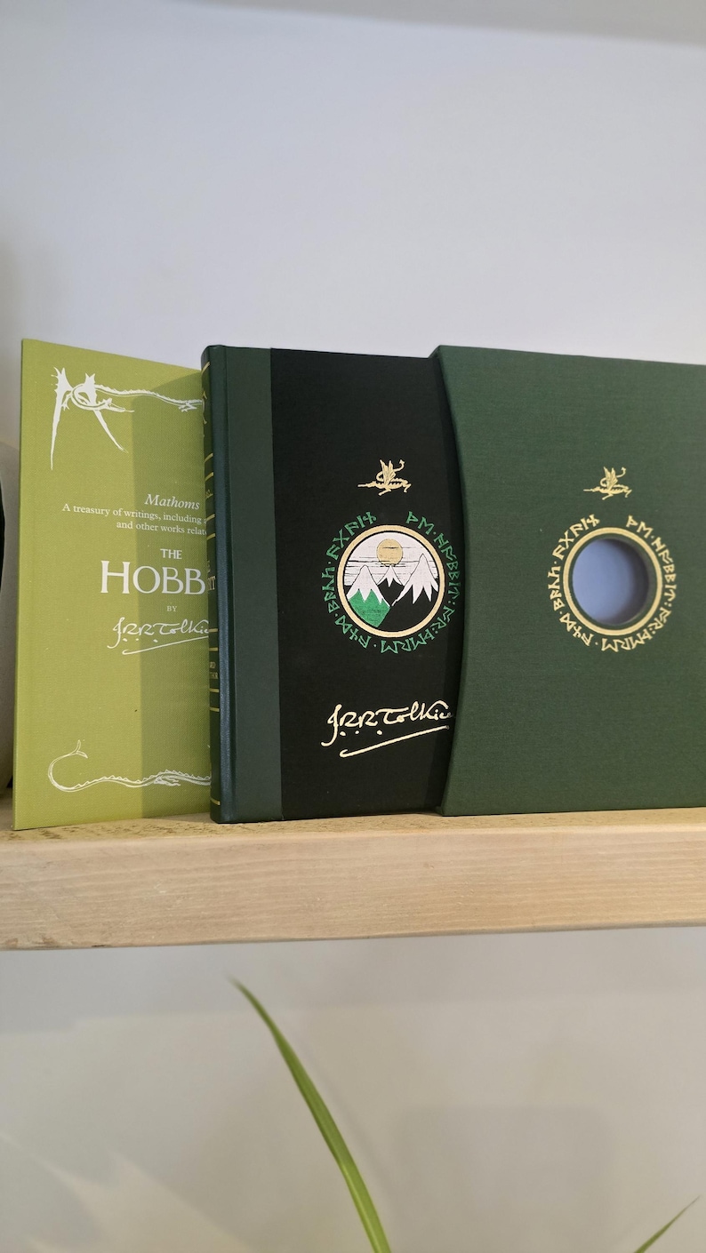 Pode incluir: Um livro de capa dura verde com o t&iacute;tulo "O Hobbit" de J.R.R. Tolkien. O livro est&aacute; em uma caixa deslizante com uma capa verde e um recorte circular que revela um fundo azul.