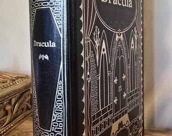 Puede incluir: Un libro de tapa dura negro titulado "Dracula" con detalles plateados. La portada y el lomo presentan ilustraciones góticas y el título. El libro está sobre una superficie de madera.