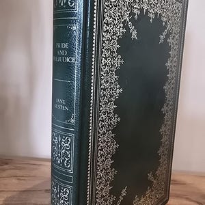 Könnte beinhalten: Ein dunkelgrünes gebundenes Buch mit dem Titel Pride and Prejudice von Jane Austen. Der Einband und der Buchrücken sind mit filigranen silbernen Mustern und dekorativen Rändern verziert. Titel und Autor stehen auf dem Buchrücken.