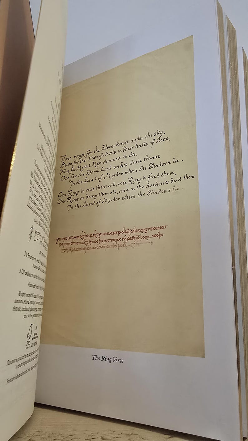 Pode incluir: Close-up de uma p&aacute;gina de um livro com uma inscri&ccedil;&atilde;o manuscrita em tinta marrom. O texto diz "Three rings for the Elven-kings under the sky, Seven for the Dwarf-lords in their halls of stone, Nine for Mortal Men doomed to die, One for the Dark Lord on his dark throne In the Land of Mordor where the Shadows lie. One Ring to rule them all, One Ring to find them, One Ring to bring them all, and in the darkness bind them In the Land of Mordor where the Shadows lie." A p&aacute;gina &eacute; intitulada "The Ring Verse".