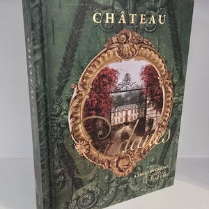 May include: A hardcover book titled "Chateau d'Ames: A True Love Story" with a green cover and gold lettering. The cover features an ornate gold frame with a picture of a chateau and red trees.