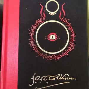 Pode incluir: Um livro preto com lombada vermelha. A capa apresenta um olho dourado com pupila vermelha dentro de um c&iacute;rculo rodeado por uma inscri&ccedil;&atilde;o dourada. A inscri&ccedil;&atilde;o diz "Um Anel para govern&aacute;-los a todos, Um Anel para encontr&aacute;-los, Um Anel para traz&ecirc;-los a todos e nas trevas prend&ecirc;-los." O livro &eacute; assinado "J.R.R. Tolkien."