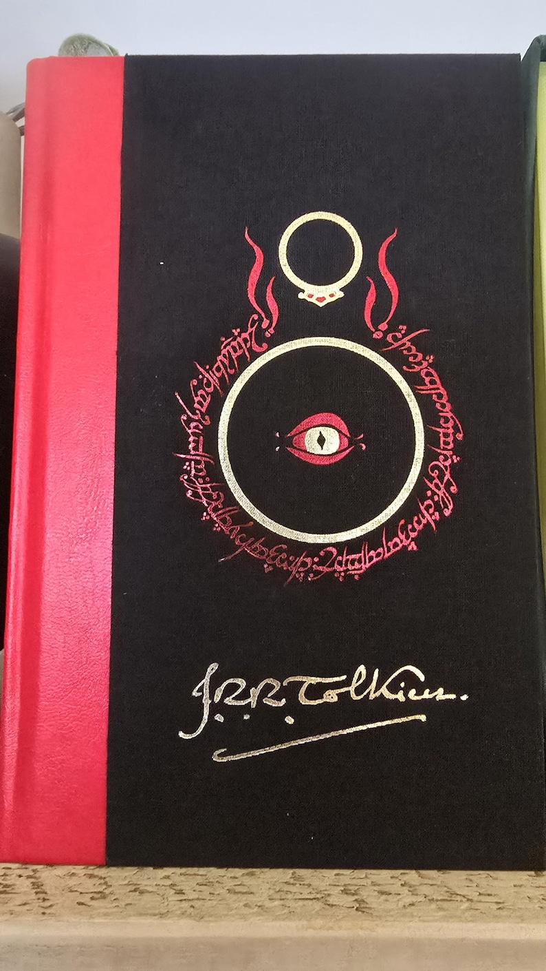 Pode incluir: Um livro preto com lombada vermelha. A capa apresenta um olho dourado com pupila vermelha dentro de um c&iacute;rculo rodeado por uma inscri&ccedil;&atilde;o dourada. A inscri&ccedil;&atilde;o diz "Um Anel para govern&aacute;-los a todos, Um Anel para encontr&aacute;-los, Um Anel para traz&ecirc;-los a todos e nas trevas prend&ecirc;-los." O livro &eacute; assinado "J.R.R. Tolkien."