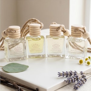May include: Four square glass car air fresheners with wooden caps and hanging cords. Scents include Eucalyptus Escape, Coco Mango, Lavender Sage, and Chamomile Eclipse. The bottles sit on a white surface with eucalyptus leaves and lavender sprigs.