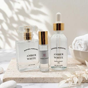 May include: Three clear glass bottles of Amber White fragrance and bath & body oil. The bottles have gold accents and are labeled with the brand name Baraka Porscha. A rolled white towel and decorative stones are also in the image.
