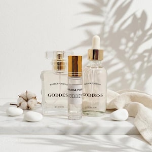 May include: Three clear glass bottles of varying sizes with gold-colored caps and labels that read "BARAKA PORSCHA GODDESS". The bottles are arranged on a white marble surface with white stones and a cotton sprig.