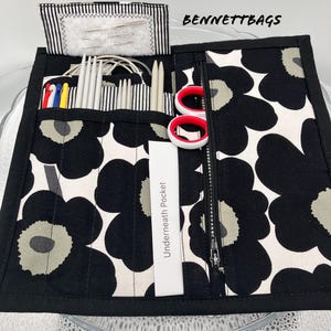 May include: Black and white fabric zippered pouch with a floral pattern. The pouch has multiple pockets for storing knitting needles, crochet hooks, and other craft supplies. The pouch also has a zippered pocket for storing scissors and other small items. The pouch is labelled "Underneath Pocket" and "BENNETT BAGS".