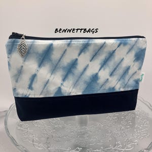 Peut inclure: Une trousse de maquillage rectangulaire avec un motif tie-dye bleu et blanc sur la moitié supérieure et un fond bleu marine uni. Le sac a une tirette de fermeture éclair en forme de feuille argentée et le texte "BENNETTBAGS".
