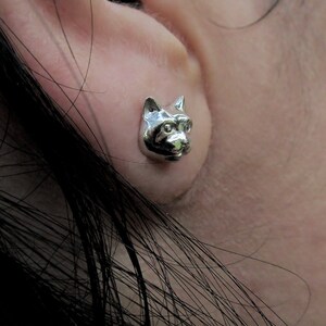 Puede incluir: Pendientes de plata con forma de cabeza de gato. La cabeza del gato está mirando hacia adelante con las orejas levantadas. Los pendientes son pequeños y delicados.