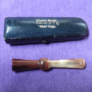 May include: A brown and white cigarette holder in a blue leather case. The case has the inscription "Captain Baylis, From the A.W.C. Motor Corps."
