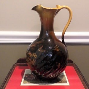 Puede incluir: Una jarra de cerámica marrón y dorada con un diseño de remolino y un motivo floral. La jarra tiene una boca ancha y un asa.