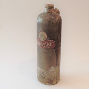 Puede incluir: Una botella de cerámica marrón con una etiqueta que dice "Nusskamp" en letras rojas y blancas. La botella tiene una pequeña asa y un tapón de corcho.