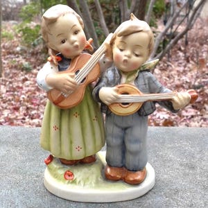 May include: A porcelain figurine of a girl and boy playing musical instruments. The girl is wearing a green dress and playing a guitar. The boy is wearing a gray jacket and playing a mandolin. The figurine is set on a white base with green grass and red flowers.