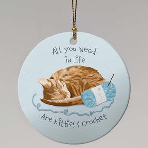 May include: A round, light blue ornament with a sleeping orange tabby cat and a blue yarn ball. Text reads "All you need in life are kitties & crochet."