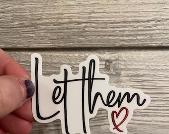 May include: A white sticker with the words "Let them" in black script, and a red heart. The sticker is held by a hand against a gray wood-grain background. The sticker is a decorative item.
